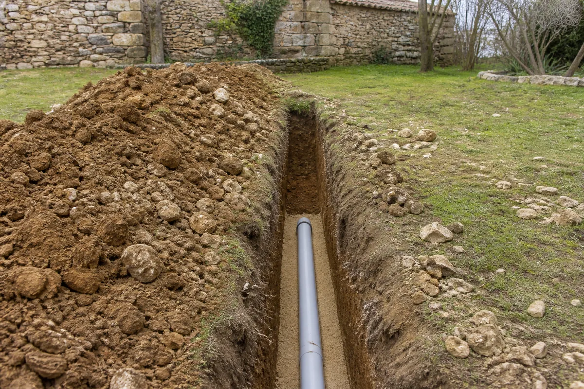 Tranchée étroite ouverte dans un jardin de campagne, tuyau d’évacuation lisse visible au fond de la fouille